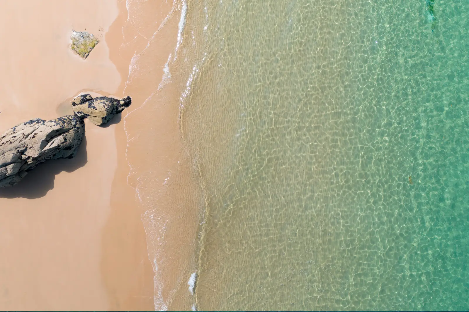 Vue aérienne d'une plage de sable clair bordée d'une eau turquoise transparente, rochers sombres posés sur l'estran