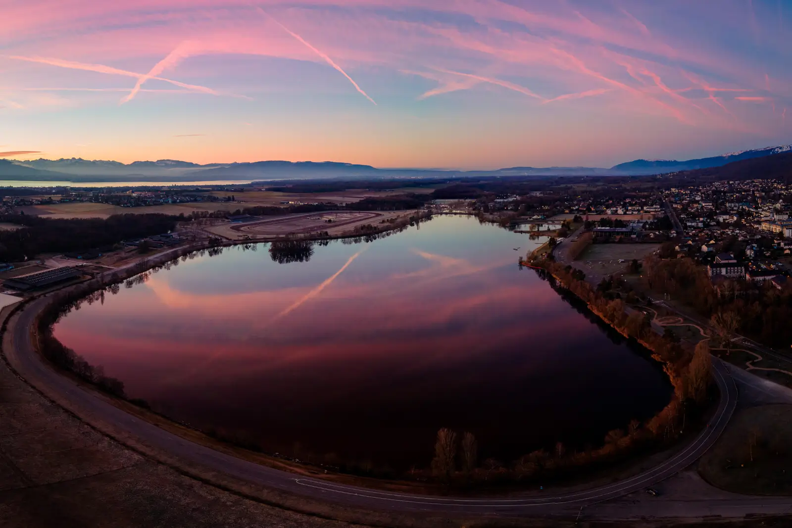 Panorama aérien d'un lac reflétant un ciel d'aube rose, entre hippodrome et village, montagnes enneigées au loin