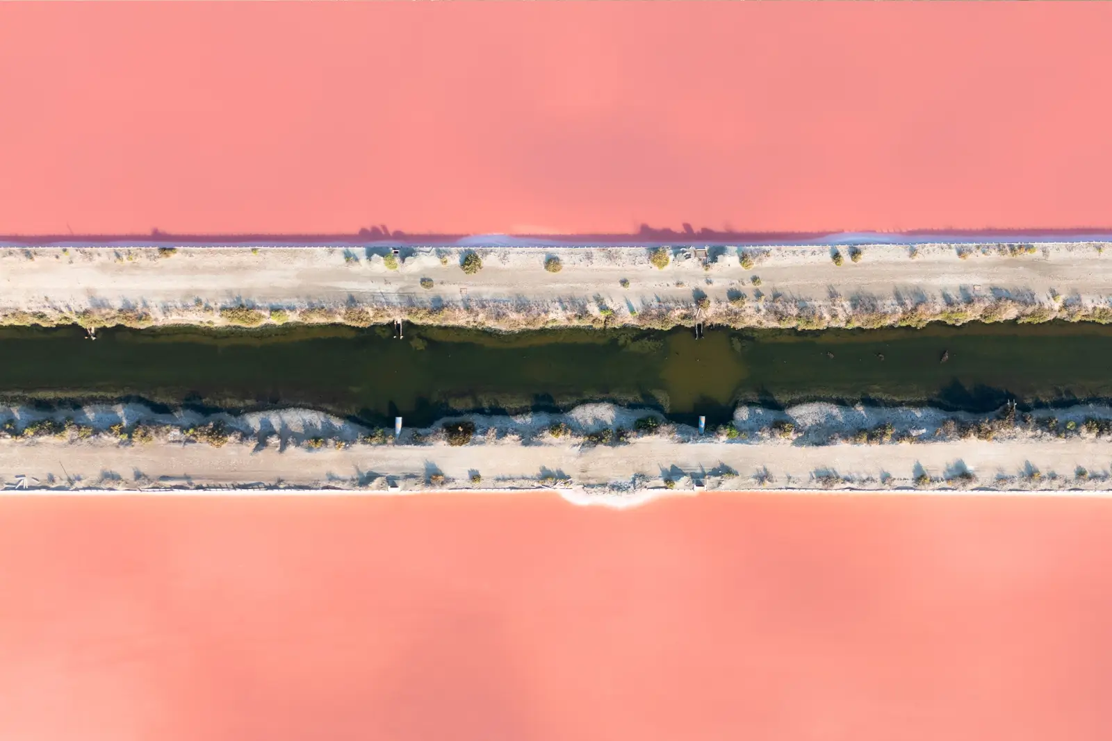 Vue aérienne de bassins de marais salants aux eaux roses, séparés par une digue de sable et un canal d'eau sombre