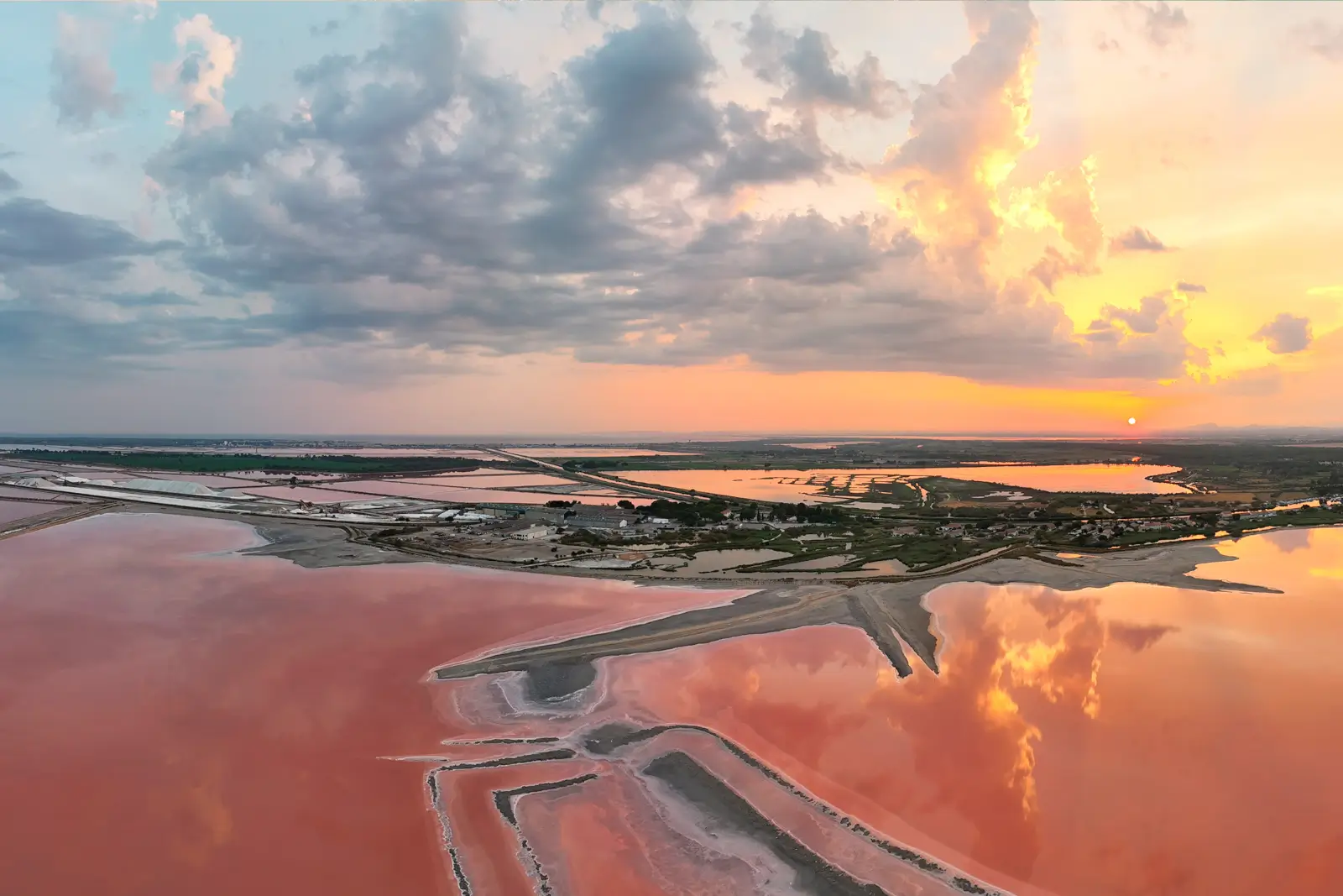 Vue aérienne de salins aux eaux roses au coucher du soleil, monticules de sel blanc et ciel flamboyant reflété dans les bassins