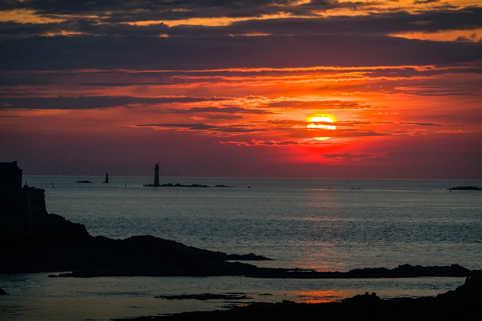 Coucher de soleil rouge et orangé sur la mer, silhouettes d'îlots et de phare au loin