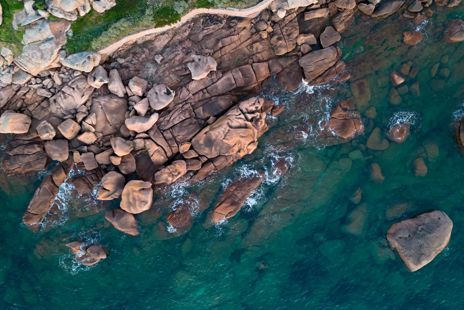 Vue aérienne de blocs de granit rose plongeant dans une mer turquoise translucide, écume légère au ras des rochers