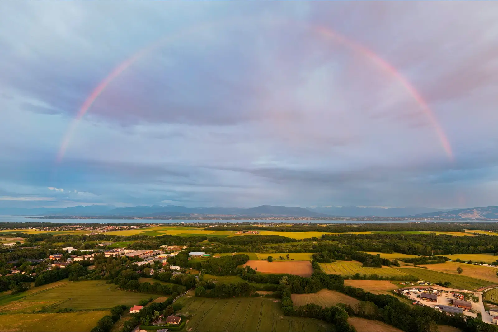 Arc-en-ciel complet au-dessus d'une plaine agricole, grand lac et montagnes à l'horizon sous un ciel rosé de fin d'orage